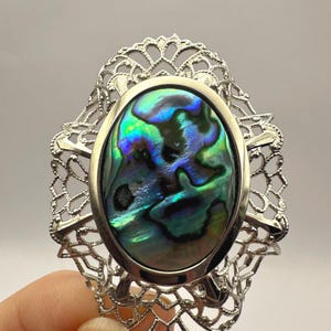 May include: A silver-toned brooch featuring an oval abalone shell with iridescent green, blue, and purple hues. The shell is set within a decorative filigree frame, creating an elegant and eye-catching accessory.