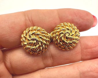 May include: A pair of gold-colored stud earrings. Each earring has a round, textured design with a woven rope-like pattern. The earrings are displayed against a light background.