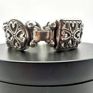 Puede incluir: Una pulsera plateada con un dise&ntilde;o ornamentado. La pulsera presenta eslabones ovalados y cuadrados, cada uno adornado con un patr&oacute;n repetido en forma de coraz&oacute;n. El cierre es visible y la pulsera se muestra sobre una superficie negra.