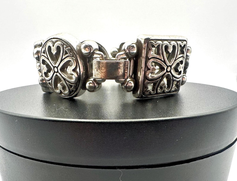 Puede incluir: Una pulsera plateada con un dise&ntilde;o ornamentado. La pulsera presenta eslabones ovalados y cuadrados, cada uno adornado con un patr&oacute;n repetido en forma de coraz&oacute;n. El cierre es visible y la pulsera se muestra sobre una superficie negra.