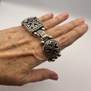 Puede incluir: Pulsera plateada con elementos decorativos cuadrados y circulares, ambos con un dise&ntilde;o en forma de coraz&oacute;n. La pulsera est&aacute; conectada por una serie de peque&ntilde;as cuentas redondas. La pulsera se lleva en una mano.