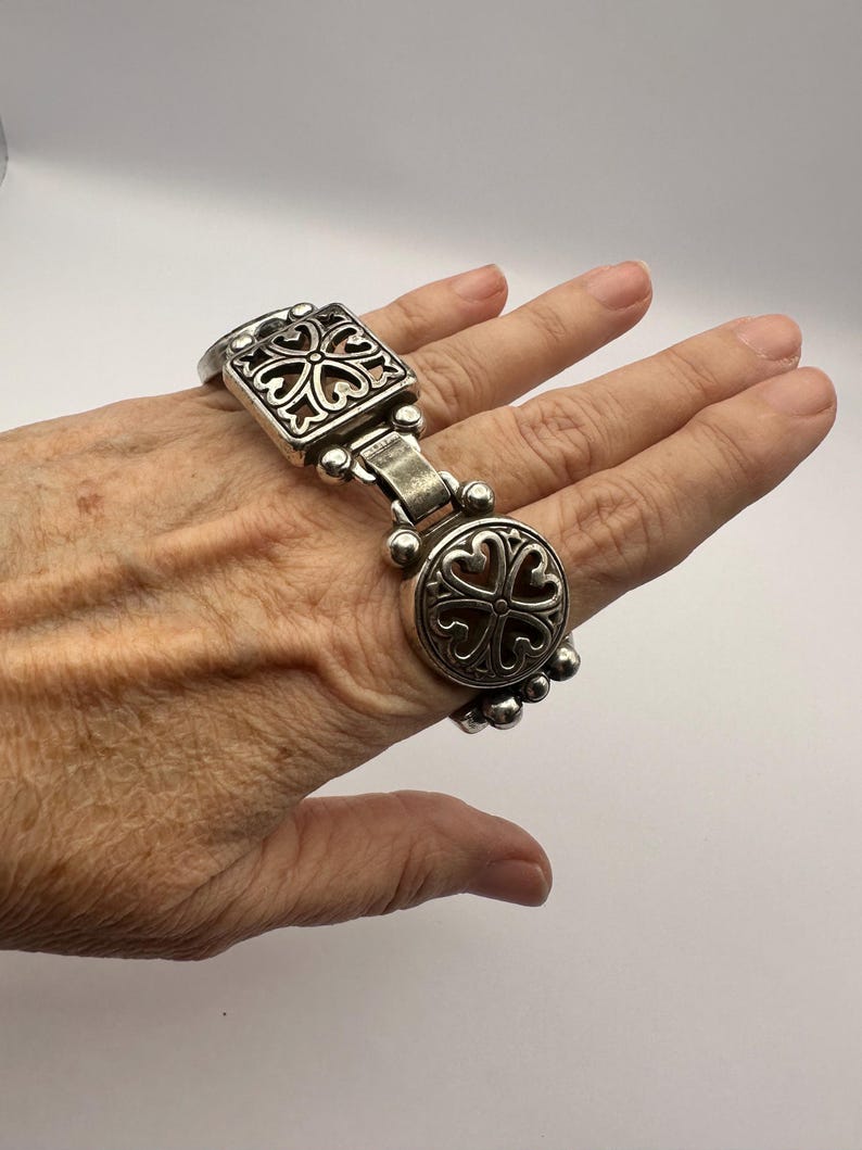 Puede incluir: Pulsera plateada con elementos decorativos cuadrados y circulares, ambos con un dise&ntilde;o en forma de coraz&oacute;n. La pulsera est&aacute; conectada por una serie de peque&ntilde;as cuentas redondas. La pulsera se lleva en una mano.