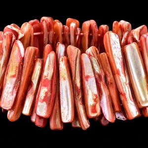 May include: A stretch bracelet composed of numerous elongated, rectangular pieces of iridescent red shell. Each piece features a smooth, polished surface with natural variations in color, ranging from deep crimson to lighter coral tones with pearlescent highlights.