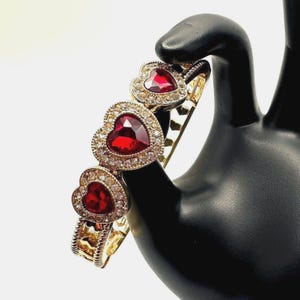 May include: Gold-toned bracelet featuring three heart-shaped red gemstones, each surrounded by smaller clear crystals. The bracelet is held by a black hand, showcasing its intricate design and sparkling details. A romantic and elegant piece of jewelry.