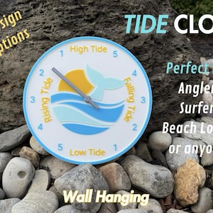 May include: A white and blue tide clock with a whale tail design. The clock face has numbers 1 through 5 and the words "High Tide", "Low Tide", "Rising Tide", and "Falling Tide". The clock is surrounded by small gray rocks and a larger rock with the words "TIDE CLOCK" in blue letters.