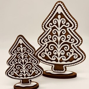 May include: Two wooden Christmas tree decorations. The trees are brown with white icing-like details, including a dotted border and intricate swirling patterns. They stand on brown bases with a touch of white at the bottom, resembling snow.