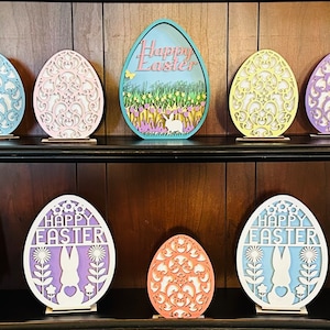 May include: A shelf display of eight wooden Easter egg decorations. The eggs are painted in various colors, including blue, pink, yellow, purple, and orange. Some of the eggs have a cut-out design, while others have the words "Happy Easter" written on them.