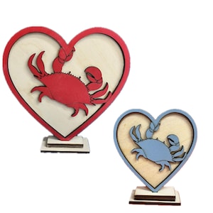 May include: Two wooden heart-shaped ornaments with a crab design. The left ornament features a red crab, while the right ornament features a blue crab.