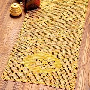 May include: A yellow crocheted table runner with a delicate pattern. The runner is on a wooden table with a sunflower in a brown vase and a honey pot.