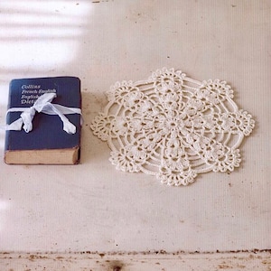 May include: A white crocheted doily with a delicate floral pattern sits on a white wooden surface next to a blue book titled "Collins French-English English-French Dictionary".