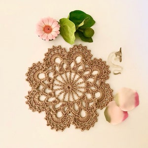 May include: A beige crocheted doily with a delicate floral pattern. The doily is surrounded by pink flower petals, a green leaf, and a white ceramic figurine.