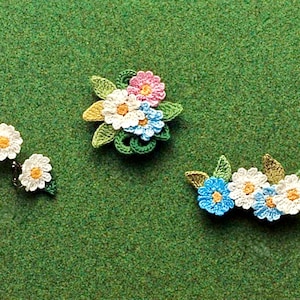 May include: Three crocheted flower embellishments with green leaves. The first embellishment is a cluster of five flowers in white, pink, and blue. The second and third embellishments are each a cluster of three flowers in white and blue.