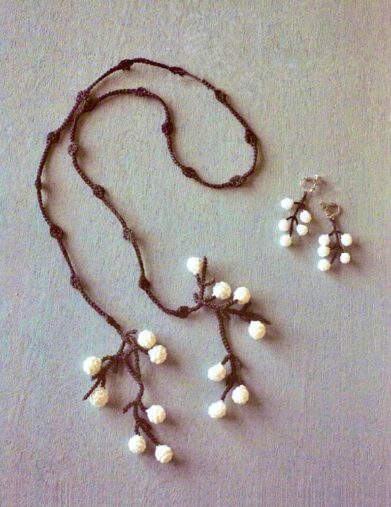 Pode incluir: Um conjunto delicado de colar e brincos de croch&ecirc; com uma corrente marrom e detalhes de flores brancas. O colar tem dois ramos com cachos de flores brancas.