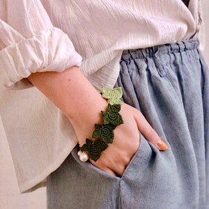 Pode incluir: Um bracelete de crochê verde com um fecho de pérola. O bracelete é feito de pequenas folhas e é usado no pulso.