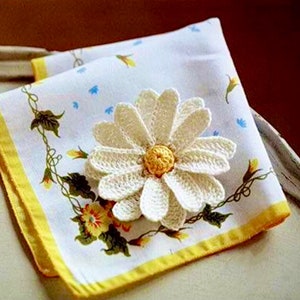 May include: A white crocheted daisy flower with a yellow center, placed on a white fabric with a yellow border and a floral pattern.