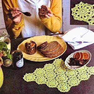 May include: A table setting with a yellow crocheted doily, a plate of bread, a small plate with cookies and dried fruit, a jar of jam, and a cup of coffee. The table is covered in a dark brown tablecloth.