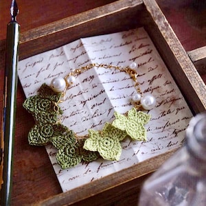 Op de afbeelding: Een delicate gouden kettingarmband met groene gecrochete bladeren en witte parel accenten. De armband wordt getoond op een stuk papier met handgeschreven tekst.