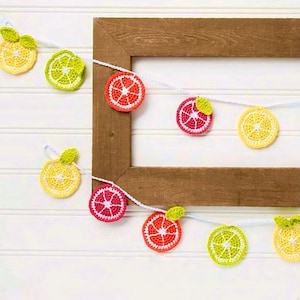 May include: A white string garland with crocheted fruit slices. The garland features alternating red, yellow, and green crocheted citrus slices.