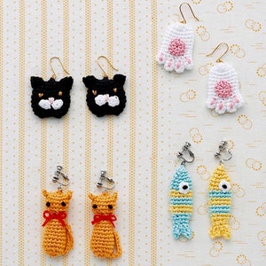Può includere: Una collezione di orecchini all'uncinetto fatti a mano in vari design. Include facce di gatto nero, impronte di zampe bianche con cuscinetti rosa, figure di gatto arancioni con fiocchi rossi e pesci blu e gialli. Ogni paio ha finiture dorate o argentate.