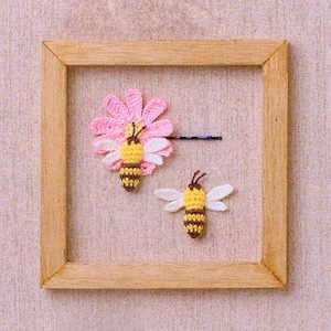 May include: Two crocheted bee hair clips, one with a pink flower, in a wooden frame.
