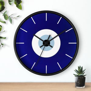 Puede incluir: Un reloj con un diseño de ojo turco azul y blanco, con manecillas negras y un marco negro. El reloj está en una pared blanca con una superficie de madera debajo.
