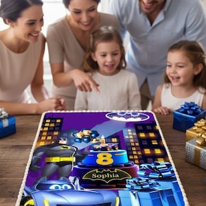 Pode incluir: Um bolo retangular com cobertura branca e borda azul. O topo apresenta um gráfico impresso do Batman, um carro de corrida azul e um bolo azul com o número 8 e o nome Sophia. A cena inclui edifícios de uma cidade.