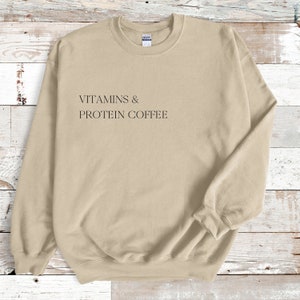 Könnte beinhalten: Ein beiger Pullover mit dem Aufdruck "VITAMINS & PROTEIN COFFEE" auf der Vorderseite.