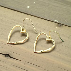 May include: A pair of gold-colored heart-shaped earrings with a hammered texture. The earrings hang from gold-colored ear wires. The earrings are displayed on a wooden surface.