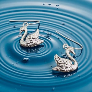 以下が含まれることがあります： 銀色の白鳥のイヤリングのペア。各イヤリングは、詳細な翼と湾曲した首を持つ白鳥が特徴です。イヤリングは、水のようなさざ波のある青い表面に表示され、反射効果を生み出しています。イヤリングの長さは約2.5cmです。