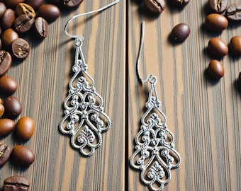 Peut inclure: Une paire de boucles d'oreilles en filigrane argenté avec un design ajouré orné. Les boucles d'oreilles ont une forme de goutte d'eau avec des détails floraux et enroulés complexes. Elles sont posées sur une surface en bois avec des grains de café éparpillés.