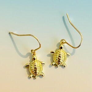 May include: A pair of gold-coloured turtle earrings. Each earring features a small, detailed turtle charm suspended from a curved ear wire. The turtles have textured shells and outstretched flippers. The earrings are displayed against a soft, gradient background.