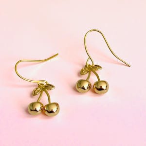 May include: A pair of gold-coloured cherry earrings. Each earring has two round, shiny cherry-shaped charms with a small leaf detail at the top, suspended from a curved ear wire. The background is a soft pink.