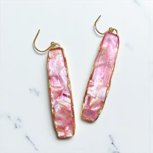 May include: A pair of rectangular earrings with a pink, iridescent mosaic design. The earrings have a gold-coloured trim and matching ear hooks. The earrings are displayed on a white marble surface.
