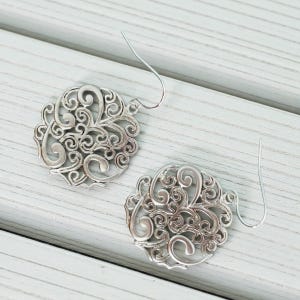 May include: Two silver-colored filigree earrings with a circular design. Each earring features an intricate pattern of swirling lines and curves, with a simple hook for the ear. The earrings are displayed on a light-colored wooden surface.