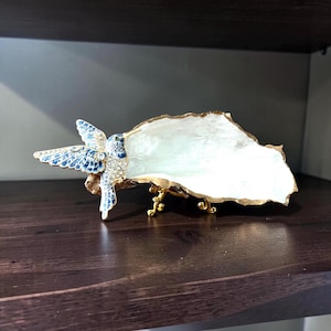 May include: A decorative oyster shell dish featuring a gold-painted rim and a gold metal stand. Attached to the side is a detailed hummingbird figurine with blue and white floral patterns, clear crystal accents, and a small green eye detail.