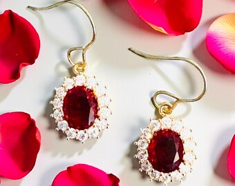 May include: A pair of gold-toned earrings with a large, oval, deep red gemstone center, surrounded by a halo of smaller, clear gemstones. The earrings are set against a backdrop of white and scattered red rose petals.
