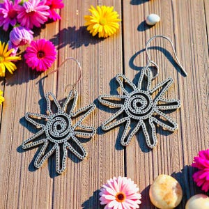 May include: A pair of silver-colored sunburst earrings with a spiral center and dotted edges. The earrings are displayed on a wooden surface, surrounded by colorful flowers.