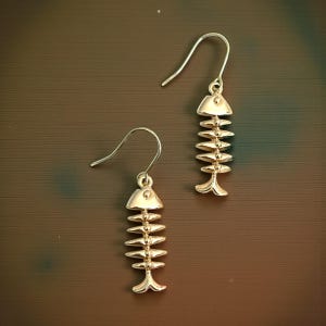 Puede incluir: Un par de pendientes dorados con forma de espina de pescado. Cada pendiente presenta un diseño de esqueleto de pez con un gancho para orejas perforadas. Los pendientes se muestran sobre un fondo marrón.