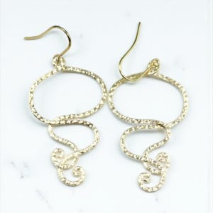 May include: A pair of gold-colored earrings with a hammered texture. Each earring features a large oval shape connected to a flowing, curved design with a small, decorative swirl at the bottom. The earrings have a hook closure.