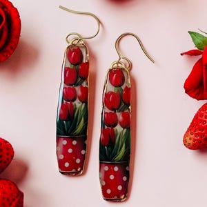 Könnte beinhalten: Ein Paar lange, rechteckige Ohrhänger mit goldfarbenen Metallrändern. Jeder Ohrring zeigt ein aufgedrucktes Motiv von roten Tulpen in einem roten Topf mit weißen Punkten. Die Ohrringe hängen an goldfarbenen Metallhaken.