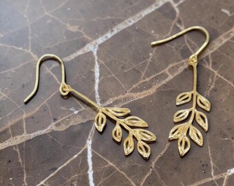 May include: A pair of gold-toned drop earrings featuring a delicate leaf branch design. Each earring consists of a curved hook wire attached to a slender stem with six open-work, teardrop-shaped leaves arranged in a symmetrical pattern along the branch.