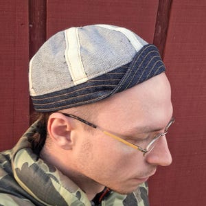 May include: A denim cap with a striped brim and vertical white panels. The cap is made of a light blue denim material and is worn by a person.
