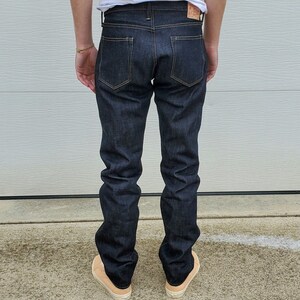 May include: Dark indigo denim jeans with a classic five-pocket design. The jeans have a straight leg fit and feature contrasting stitching. A leather patch is visible on the back waistband. The person is wearing beige sneakers.