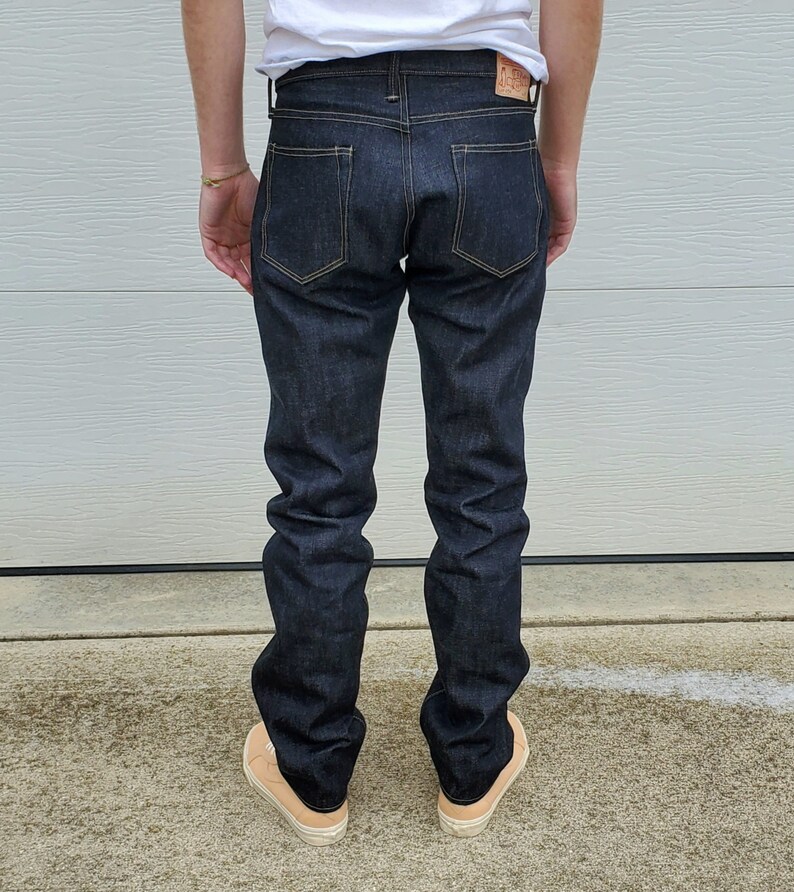 May include: Dark indigo denim jeans with a classic five-pocket design. The jeans have a straight leg fit and feature contrasting stitching. A leather patch is visible on the back waistband. The person is wearing beige sneakers.