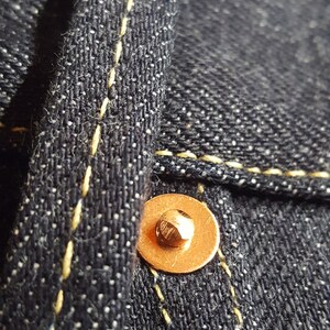 May include: Close-up of dark blue denim fabric with yellow stitching and a copper-colored rivet. The texture of the denim is visible, with small white flecks adding detail. The stitching is neat and precise.