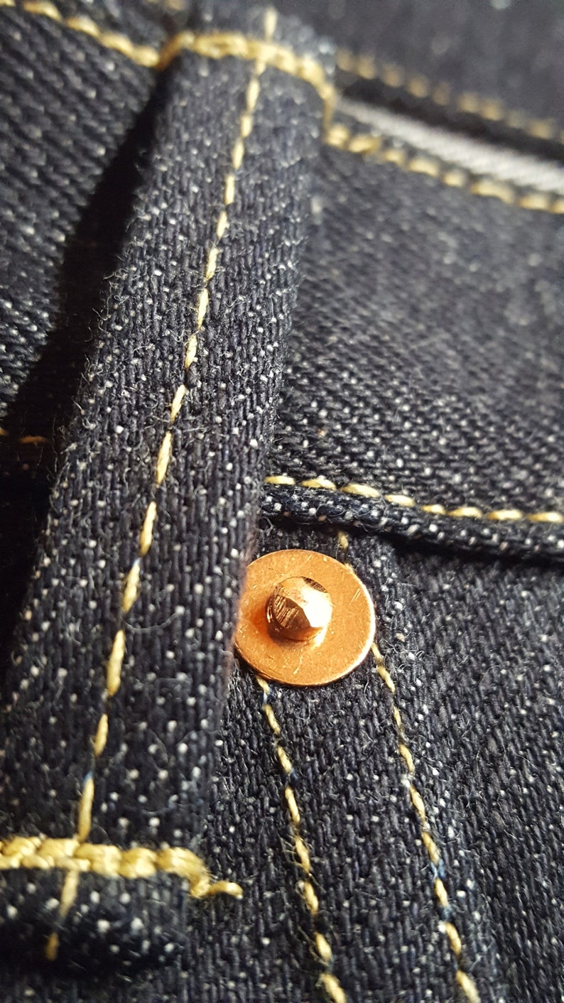 May include: Close-up of dark blue denim fabric with yellow stitching and a copper-colored rivet. The texture of the denim is visible, with small white flecks adding detail. The stitching is neat and precise.