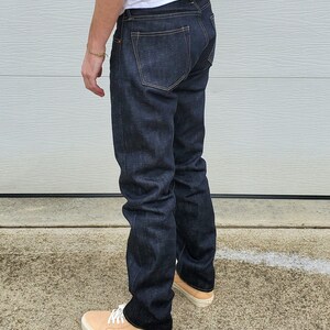 May include: Dark wash denim jeans with a classic five-pocket design. The jeans have a straight leg fit and are worn with light tan sneakers with white laces. The jeans are a deep indigo color.