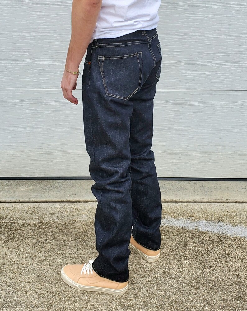 May include: Dark wash denim jeans with a classic five-pocket design. The jeans have a straight leg fit and are worn with light tan sneakers with white laces. The jeans are a deep indigo color.