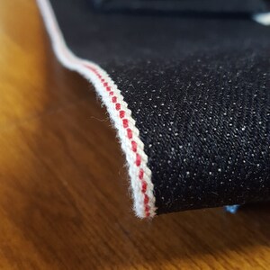 May include: Close-up of dark blue denim fabric with a white and red selvedge edge. The fabric is folded, revealing the texture and color of the denim. The selvedge edge is a clean, finished edge.