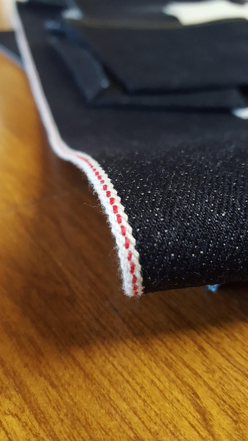 May include: Close-up of dark blue denim fabric with a white and red selvedge edge. The fabric is folded, revealing the texture and color of the denim. The selvedge edge is a clean, finished edge.